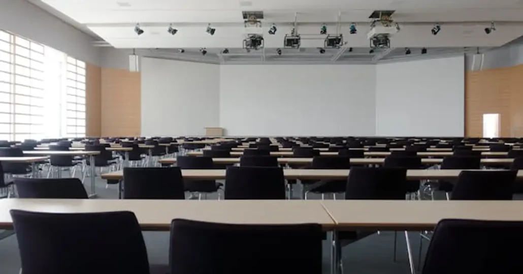 An empty conference room with rows of tables and black chairs facing a large projection screen. The room is well-lit with natural light from large windows on the left side. Ceiling-mounted projectors are visible.