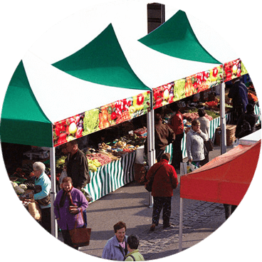 An outdoor market scene with people browsing stalls under green and white canopy tents. The stalls display a variety of fresh fruits and vegetables. Shoppers walk and interact in the sunlit area.