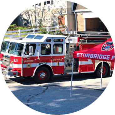 A red fire truck parked on a street near a red Sturbridge Fire Department tent. The fire truck has Engine 1 written on its side. Its a sunny day with shadows on the ground.