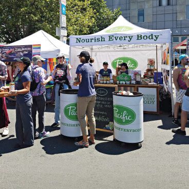 People gather around a Nourish Every Body booth at an outdoor event. The booth is set up with a canopy and has branded displays and signs. A few attendees are interacting with staff members.