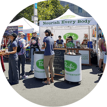 People gather at an outdoor market booth with a white tent that reads Nourish Every Body. Two promotional stands display the Pacific logo. Trees and other colorful booths are in the background.