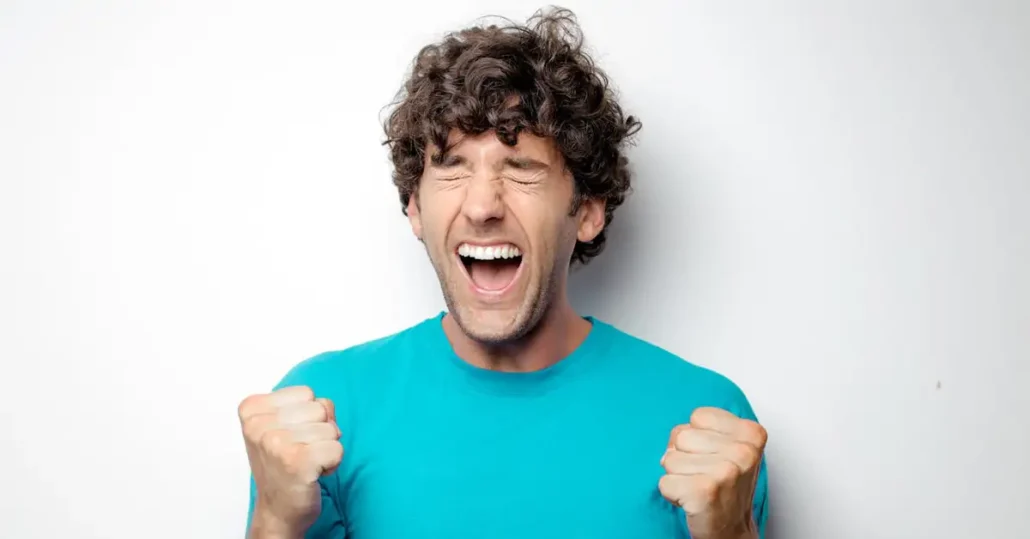 A person with curly hair wearing a bright blue shirt is exuberantly expressing joy. Their eyes are closed, mouth open, and fists clenched in excitement against a plain white background.