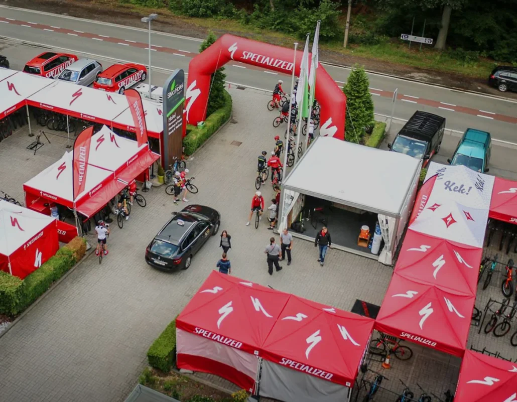 Aerial view of a Specialized cycling event with red tents and banners, people gathered around bikes, a few cars parked nearby, and trees in the background. An inflatable archway marks the event entrance.