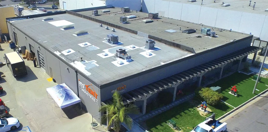 Aerial view of a large industrial building with a flat roof. The building has the logo inTENT on the side. There is a small garden area with people near the entrance. Several air conditioning units are visible on the roof.
