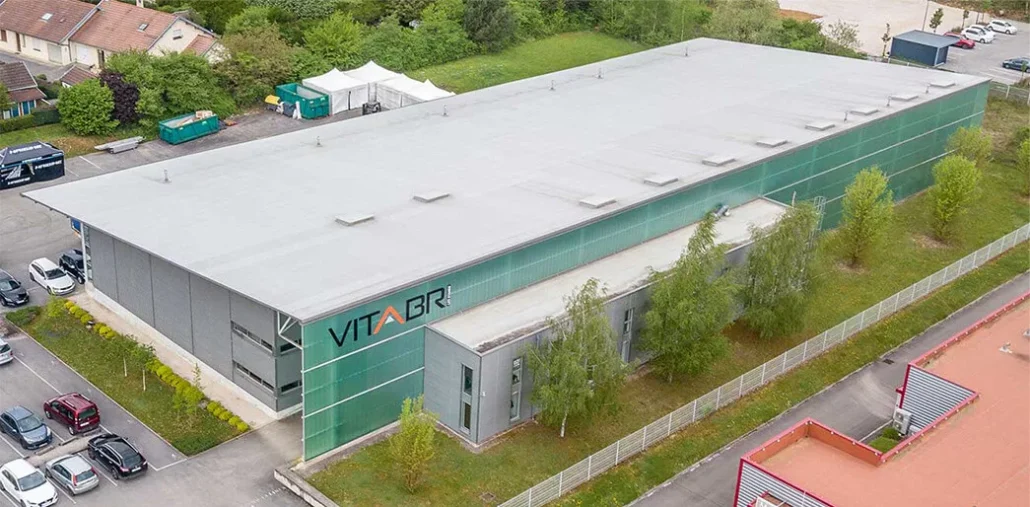 An aerial view of a modern, rectangular building with a silver roof and green-tinted glass walls. The building is labeled VITA BR. Its surrounded by trees, a parking area, and adjacent structures.