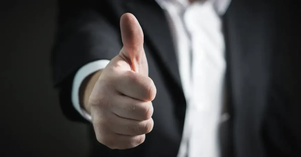 A person in a dark suit and white shirt giving a thumbs-up gesture against a dark background. The focus is on the hand, with the thumb prominently displayed in the foreground.
