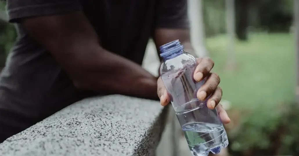 Person holding a partially filled plastic water bottle while leaning on a textured stone surface. The background is blurred with greenery, suggesting an outdoor setting.