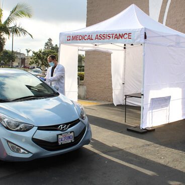 A medical professional in a white coat stands outside a white tent labeled Medical Assistance near a parked blue Hyundai. The setup is in an outdoor area with palm trees visible in the background.