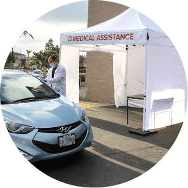 A healthcare worker in a white coat and mask stands beside a blue car under a white tent labeled Medical Assistance in a parking lot.