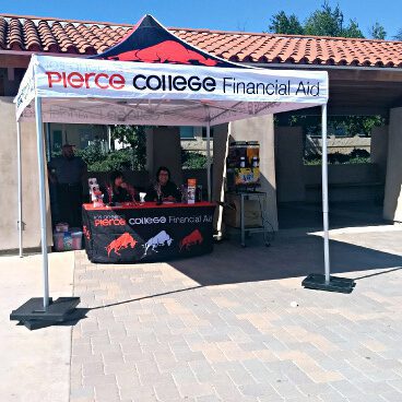 A booth with a tent labeled Pierce College Financial Aid is set up outdoors. Two people sit behind a table covered with promotional materials and flyers. Nearby, a stack of boxes is visible. The area is sunny with a tiled roof in the background.