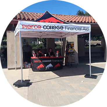 A booth under a canopy labeled Pierce College Financial Aid. The booth has people sitting behind a table with brochures and giveaways. The setup is outdoors in a sunny area with trees in the background.