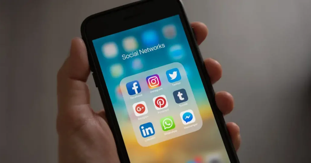 A hand holds a smartphone displaying a Social Networks folder on the screen. The folder contains icons for Facebook, Instagram, Twitter, Google+, Pinterest, Tumblr, LinkedIn, WhatsApp, and Messenger against a blurred background.
