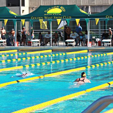 Outdoor swimming competition at Golden West Swim Club. Several swimmers in lanes with onlookers seated under green tents. Bright, sunny day with yellow lane dividers in the pool.