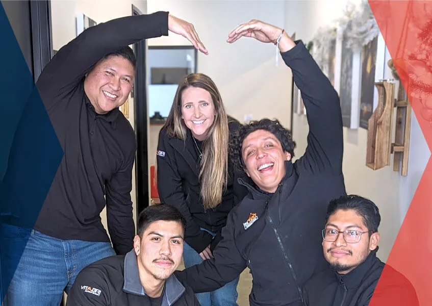 A group of five people posing together indoors, forming the letters V, I, P, E, and R with their arms. They are all smiling and wearing black jackets. The background features a hallway with hanging decorations.