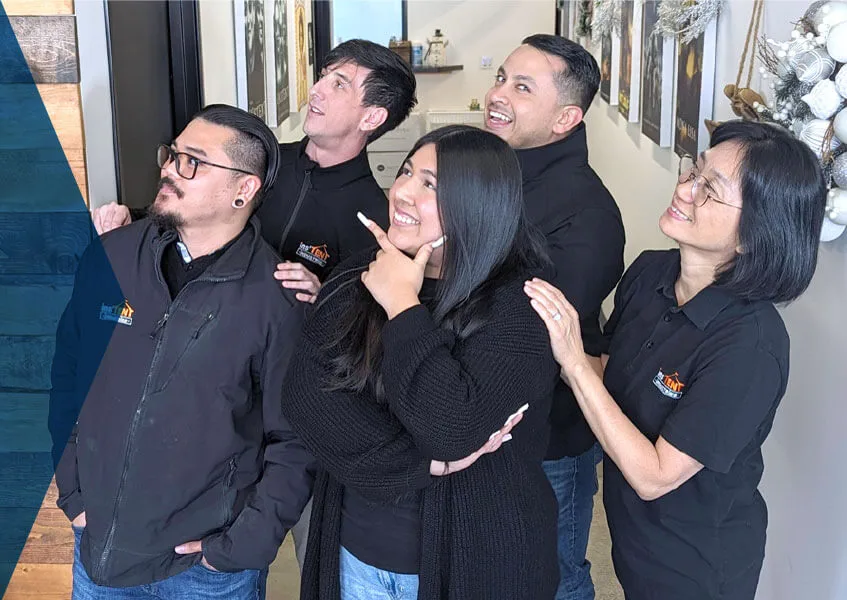 A group of five people, wearing black jackets with logos, stand indoors and look upwards with smiles and various playful expressions. They are positioned in a hallway with art on the wall and some decorative plants.