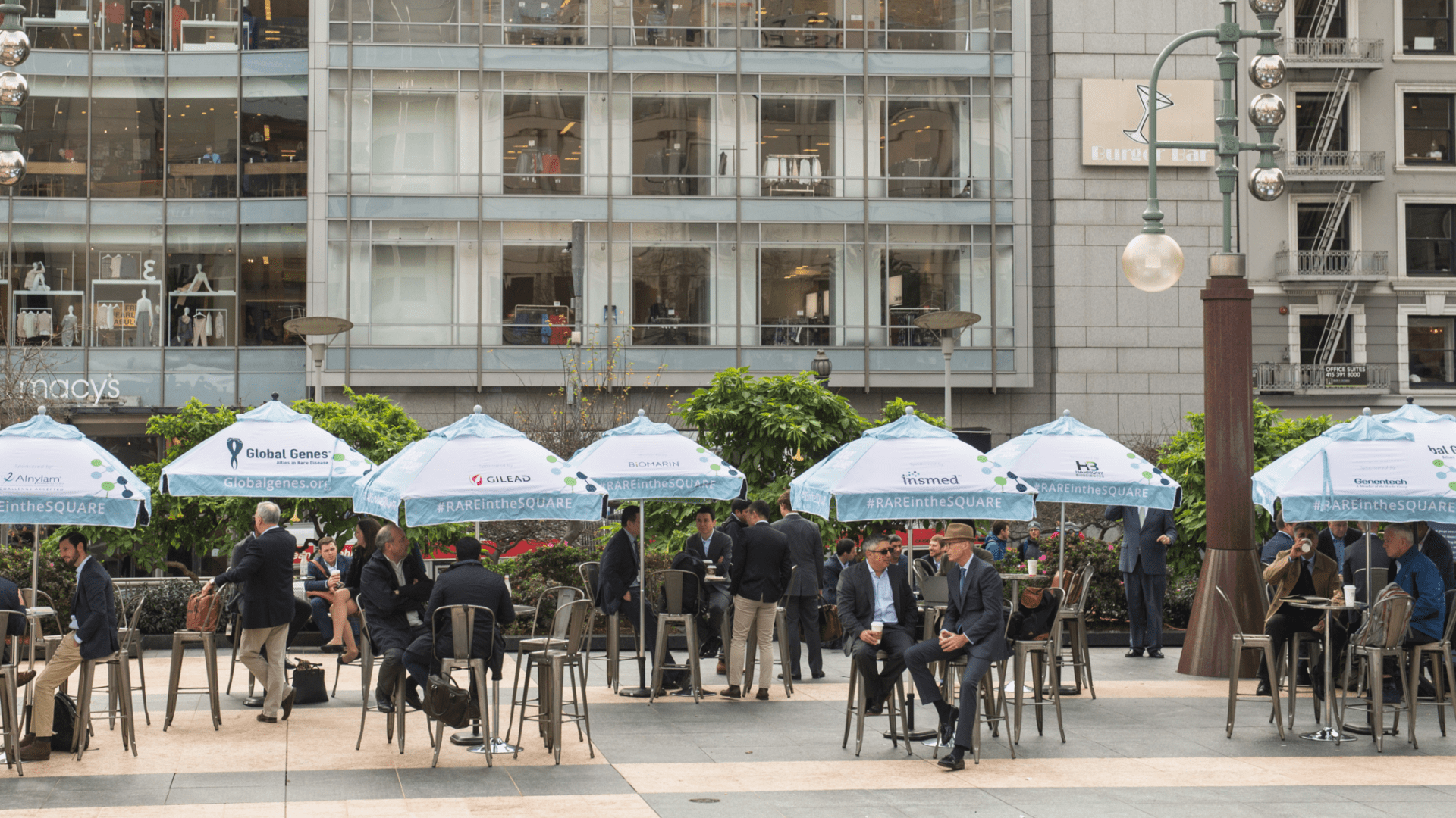 People are seated at tables under branded umbrellas in an outdoor area near a tall building with large windows. The setting appears to be a public square with businesses visible in the background.