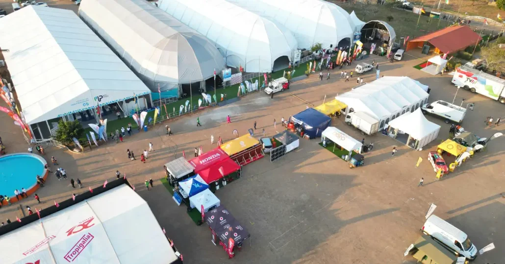 Aerial view of an outdoor event showcasing large white tents, vendor booths with colorful canopies, and people walking between setups. Vehicles are parked nearby, and a small circular pool is visible in the corner.