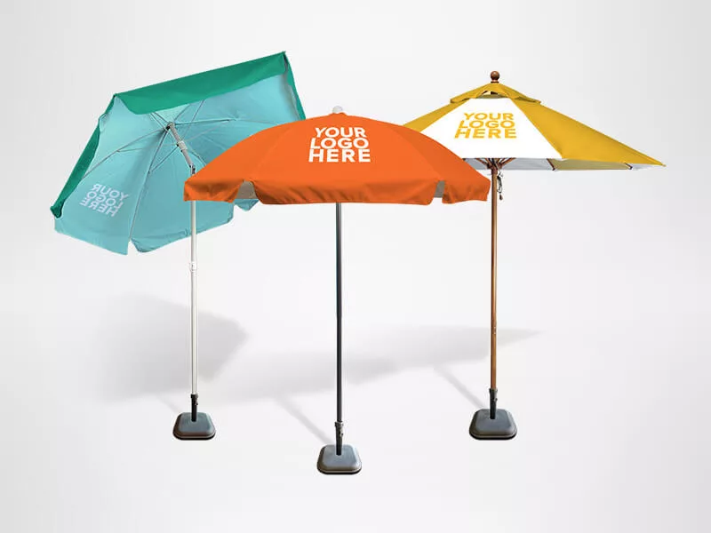 Three promotional umbrellas with placeholders for logos. One umbrella is green, another is orange, and the third is yellow and white. They have sturdy bases and are set against a plain light background.