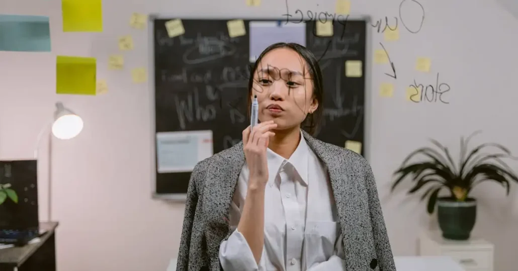 A person in a formal outfit stands in front of a glass wall covered in writing and sticky notes. They appear thoughtful, with one hand on their chin. Behind them, a blackboard with more notes is visible, along with a lamp and a potted plant.