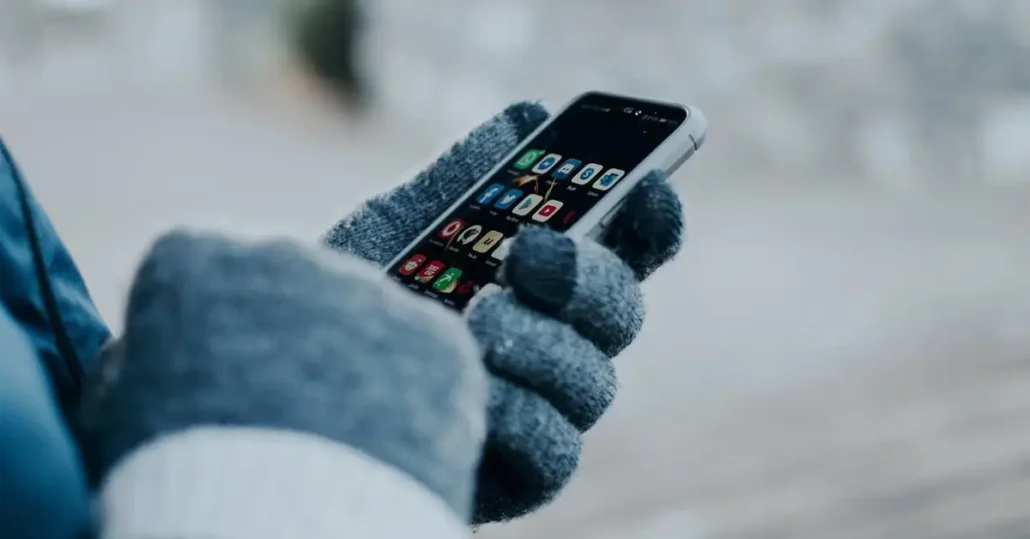 A person wearing gray gloves holds a smartphone with various app icons displayed on the screen, including messages and social media apps. The background is blurred, suggesting the scene is outdoors.