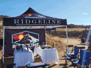 A Ridgeline-branded outdoor tent set up in a sunny, grassy area with two tables covered in white cloth, holding various gear. Blue folding chairs are arranged nearby, and a ridgeline logo with mountains and sun is visible on the tent.