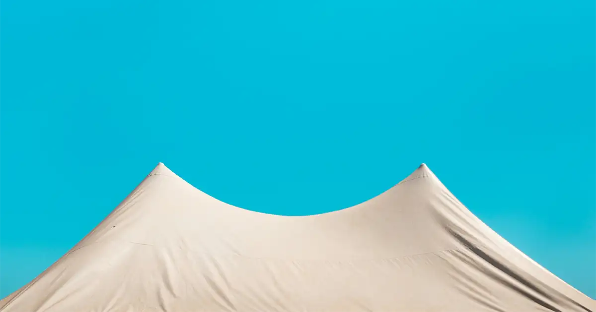 A beige tent with pointed peaks stands against a bright blue sky, creating a minimalistic and contrasting scene.