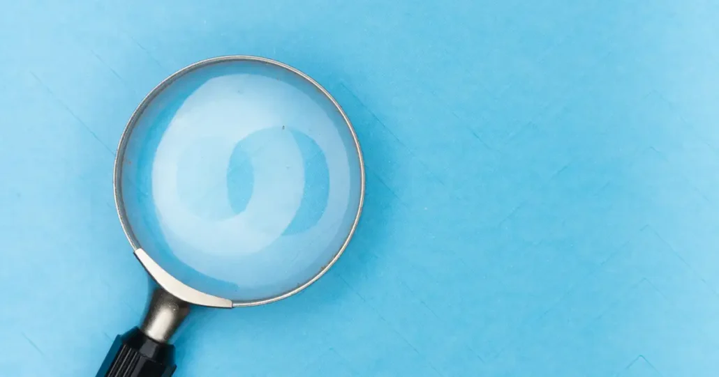 A magnifying glass with a black handle is positioned on the left side of a blue textured background, casting a shadow. The glass lens reflects the pattern, creating a visible magnified circle. The right side of the image has open space.