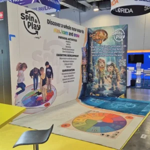 Exhibit booth featuring Spin & Play educational games. Displays include a colorful game mat on the floor and a backdrop with promotional graphics about physical activities, cognitive development, and gamification. Two children shown playing on the mat.