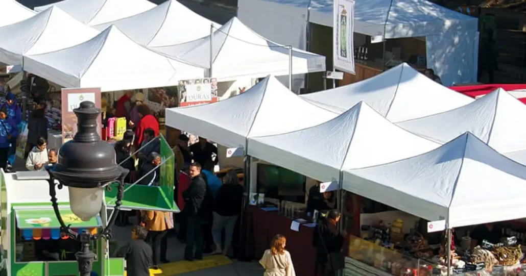 A bustling outdoor market with white tented stalls. People gather around browsing various goods, including food and crafts. A black street lamp is in the foreground, and there are trees casting shadows on the scene.