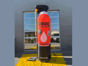 A large inflatable can shaped like a Kroil penetrant aerosol can stands outdoors on a yellow tarp. The label reads Aerokroil with details about performance and origin. The background features a building with reflective windows.