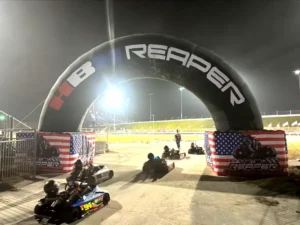 Go-kart racers drive under a large inflatable arch labeled Reaper at night. The path is illuminated, and American flags are displayed on either side of the arch. The track is surrounded by bright lights.