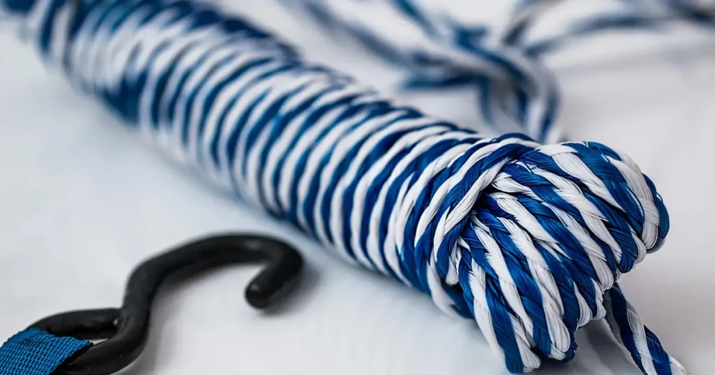 A close-up of a coiled blue and white striped rope with an attached black hook resting on a white surface. The rope appears tightly wound and the hook is slightly curved.