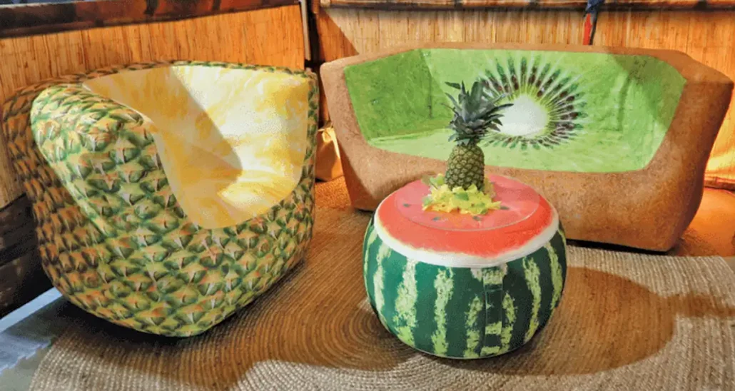 Inflatable custom furniture set featuring a pineapple-inspired chair, a kiwi-themed couch, and a watermelon-shaped table with a pineapple top, all set on a woven mat background.