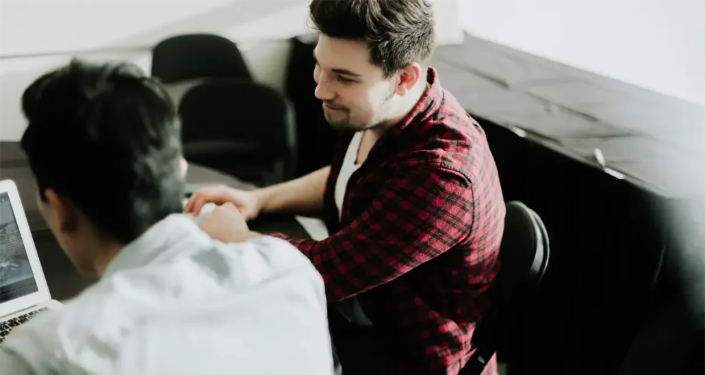 Two people are seated at a table, engaged in conversation. One person with dark hair is wearing a red plaid shirt, and the other has dark hair and is wearing a light shirt. A laptop is open in front of them. The setting appears to be a casual meeting.