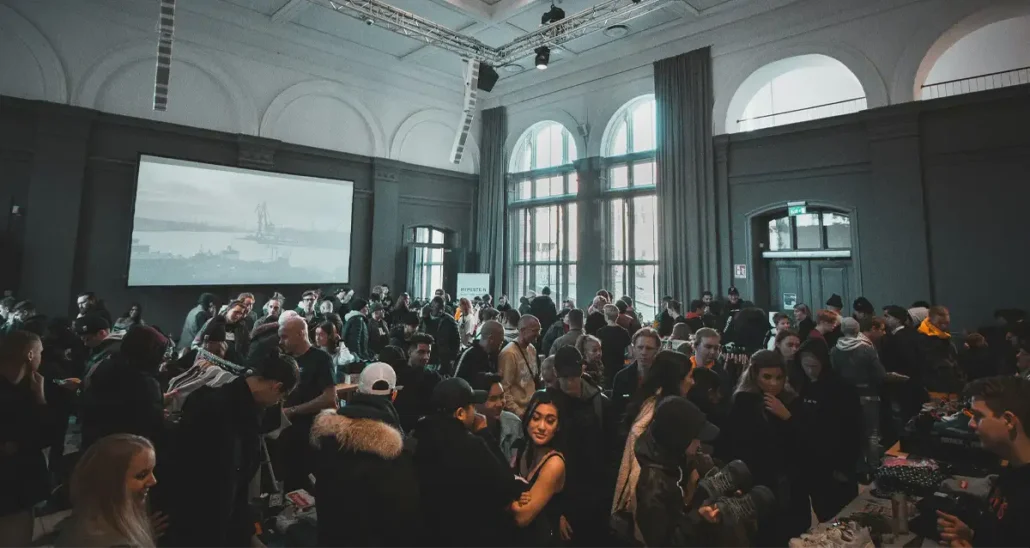A large crowd of people in a spacious hall with arched windows and high ceilings. The group appears to be interacting, browsing items on tables, and socializing. A large screen in the background displays an image or presentation.