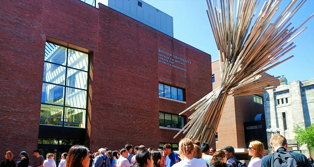 A group of people stand outside a brick building with Boston University School of Law written on it. A large abstract metal sculpture is prominently featured to the right. The scene is bright and sunny.