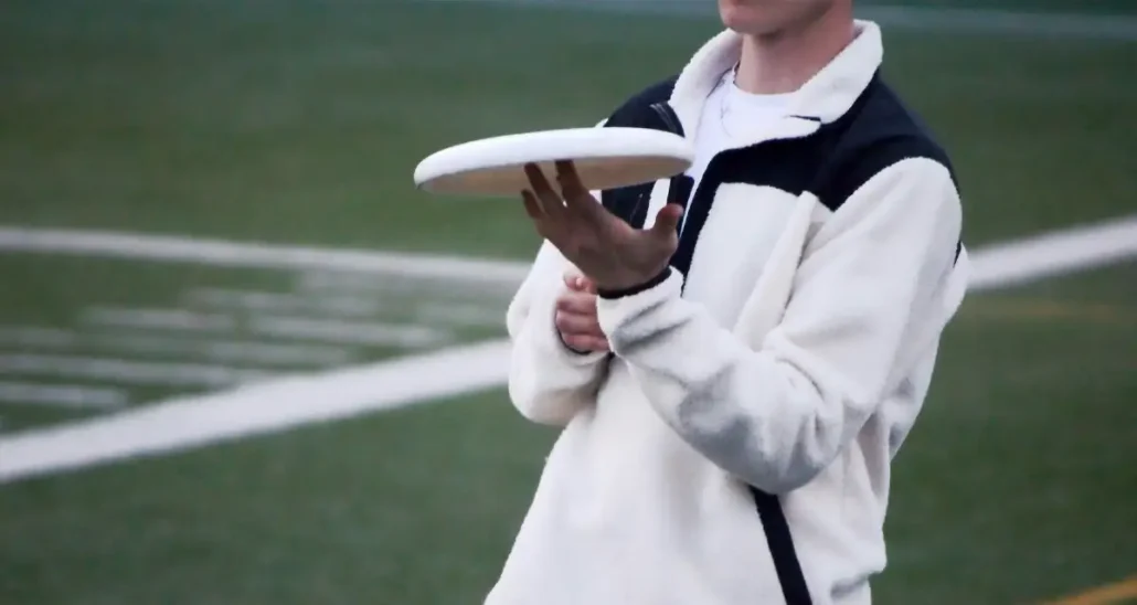 A person in a white and black fleece jacket holds a white frisbee with one hand, standing on a sports field with green artificial turf and white lines in the background.