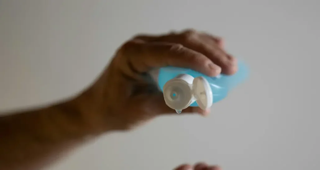 A person holding a bottle of blue hand sanitizer, with a droplet falling from the nozzle. The background is plain and blurred, highlighting the focus on hand hygiene.