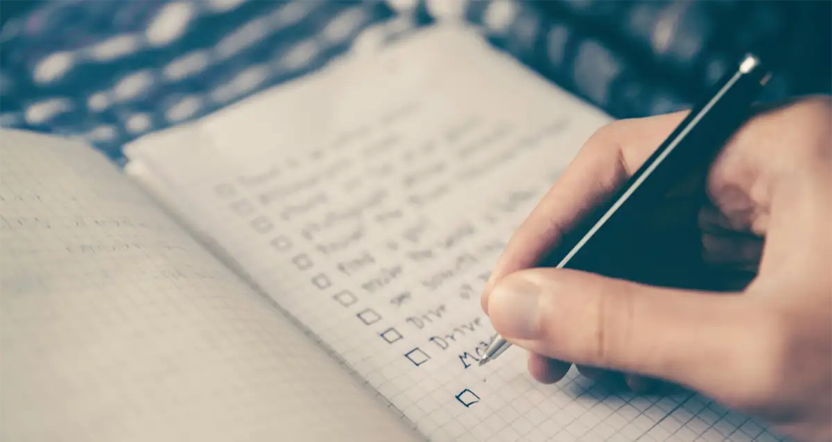 A hand holding a pen is completing an event budget checklist on graph paper. The list items are partially visible, with checkboxes next to each line. The background is blurred, focusing attention on the meticulous writing task.