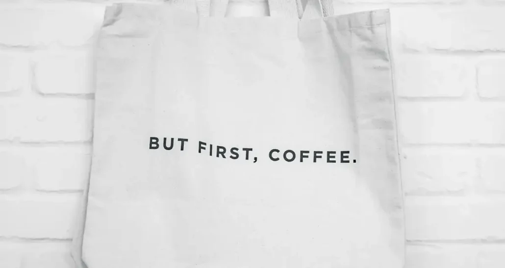 A white tote bag hangs against a white brick wall. The bag features the text BUT FIRST, COFFEE. in bold black letters.