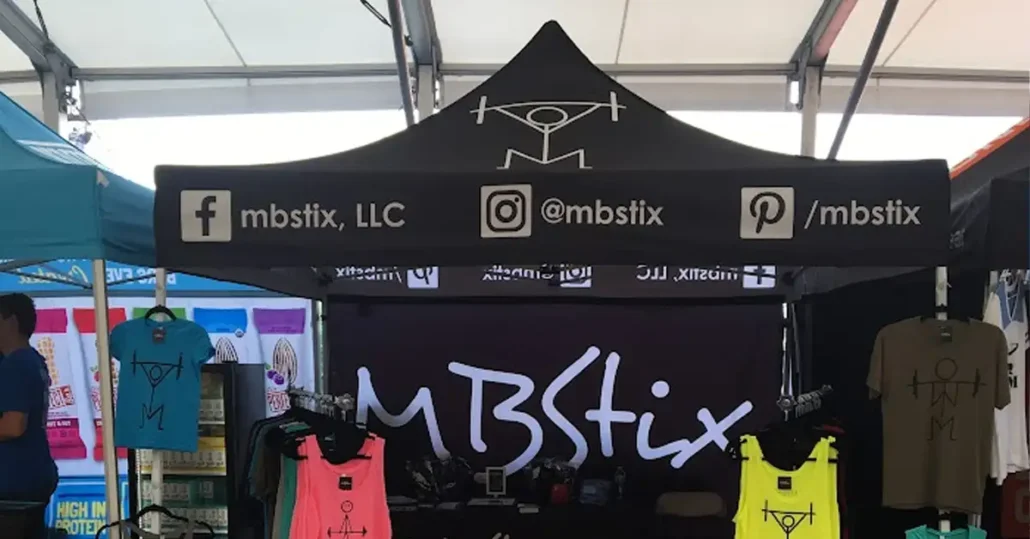 A booth with a black canopy displaying social media handles for mbstix. Underneath, various colorful tank tops with the MBS logo are showcased. The setup is at an outdoor market or festival.