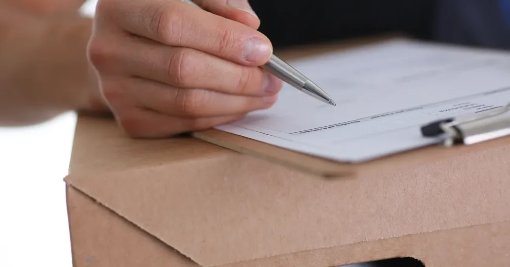 A hand holding a pen is poised above a paper on a clipboard. The clipboard rests on a cardboard box, indicating the person might be filling out a form or checklist, possibly related to shipping or delivery.