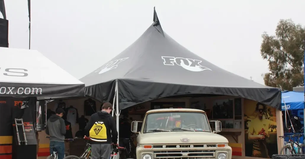 A vintage truck is parked under a large tent with the Fox logo on top. People are gathered around, some holding bikes. The tent is part of an outdoor exhibition with various items on display.
