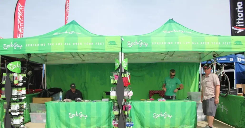 Two green SockGuy tents at an outdoor event showcase a variety of colorful socks on display racks. Three people are present: two inside the booth and one walking nearby. Banners wave in the background under a clear sky.