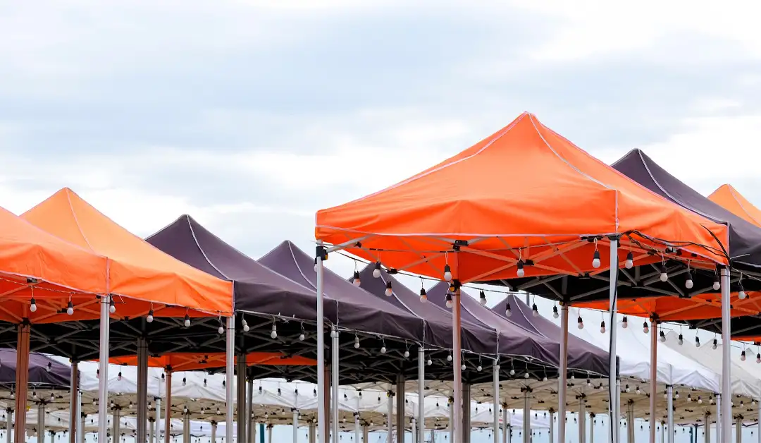 Rows of outdoor market stalls with orange and black canopies. Strings of lights are attached underneath. The sky is overcast.