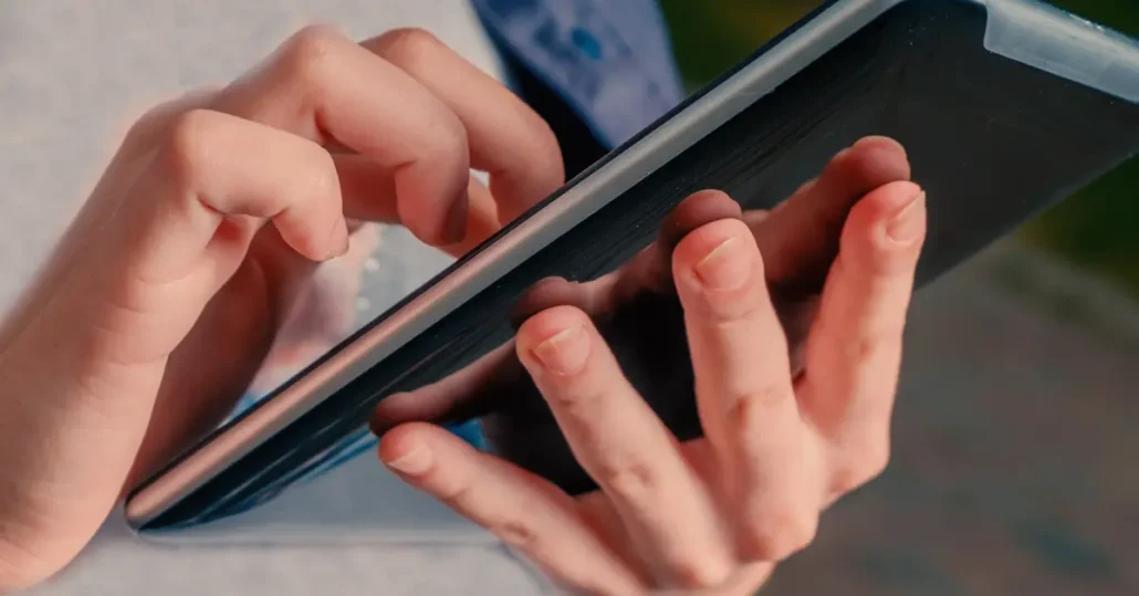 Two hands are holding and interacting with a tablet device. One hand is gripping the device from the side, while the other hands fingers are poised over the screen, suggesting typing or swiping. The background is blurred outdoors.