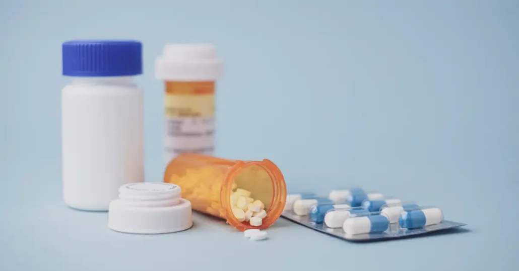 Close-up of various medication containers: a white bottle with a blue cap, a small white jar, a pill bottle with a few pills spilled, an amber prescription bottle with a label, and a blister pack of blue and white capsules. Blue background.