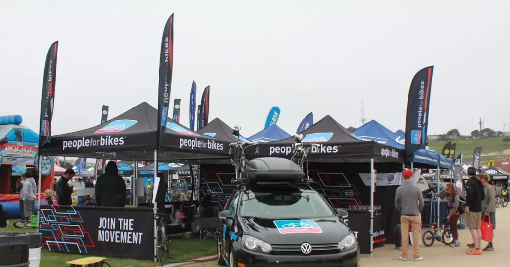 A cycling event features multiple black tents with People for Bikes branding. A black vehicle with bike racks is parked in front, surrounded by people and various event stalls under a cloudy sky.