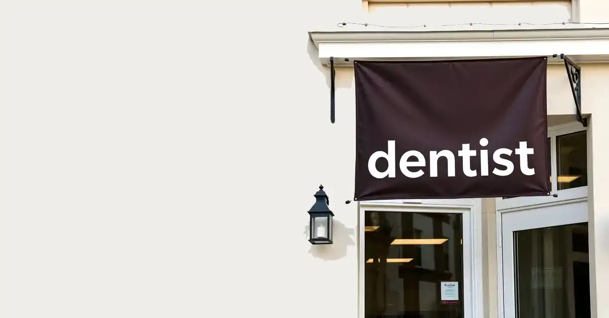 A building facade featuring a dark sign with the word dentist in white. Below the sign is a glass door and window, and a black lantern-style light fixture on the wall.