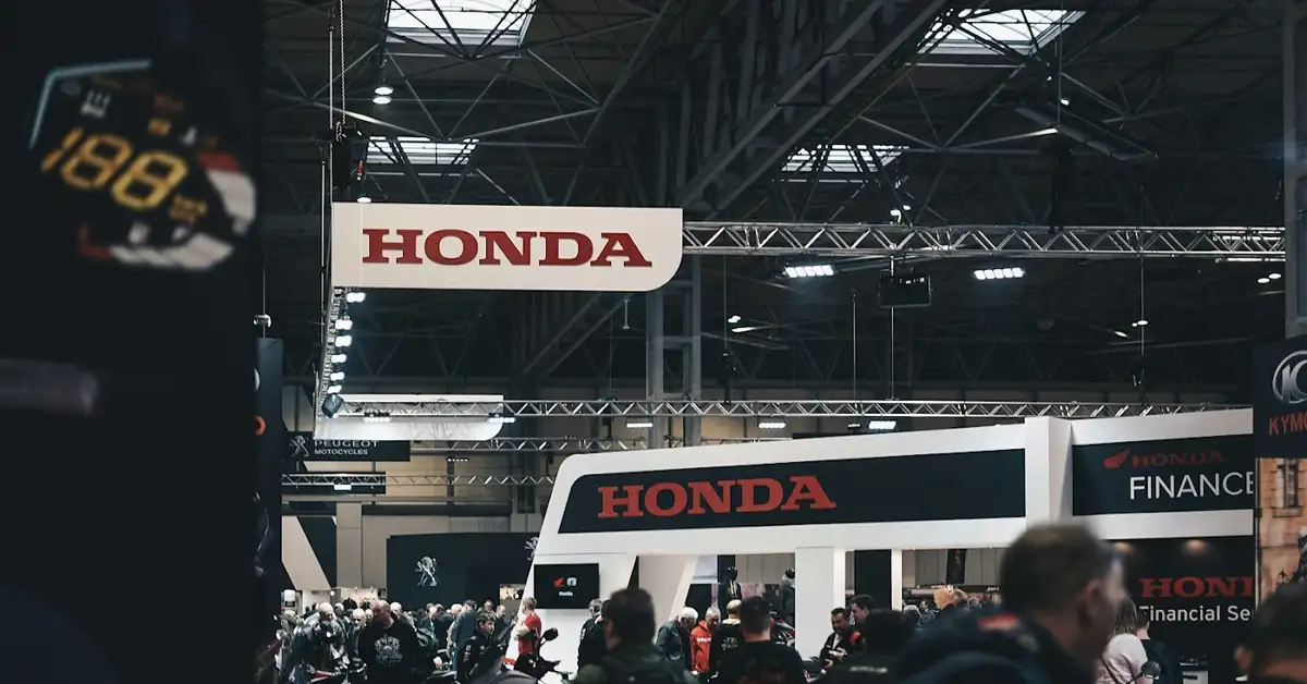 A busy automotive exhibition hall features prominent Honda banners and displays. Visitors walk around various stalls, with the Honda logo prominently visible above. The hall has a high ceiling with visible lighting and structural elements.
